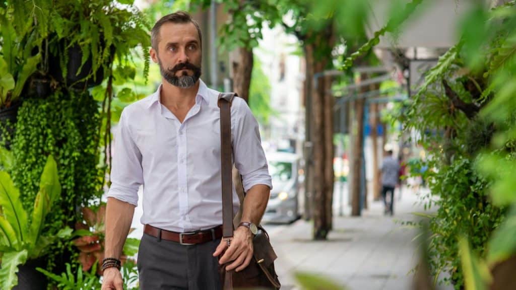 A stylish man with a beard and mustache walks on a tree-lined street.