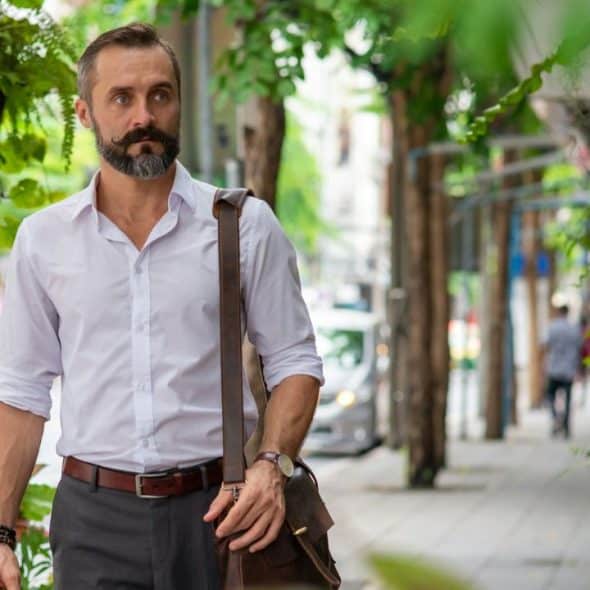 A stylish man with a beard and mustache walks on a tree-lined street.