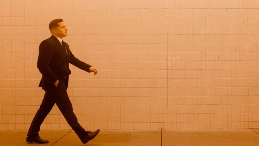 A man in a suit walks confidently with his hands in his pockets, past a textured wall.