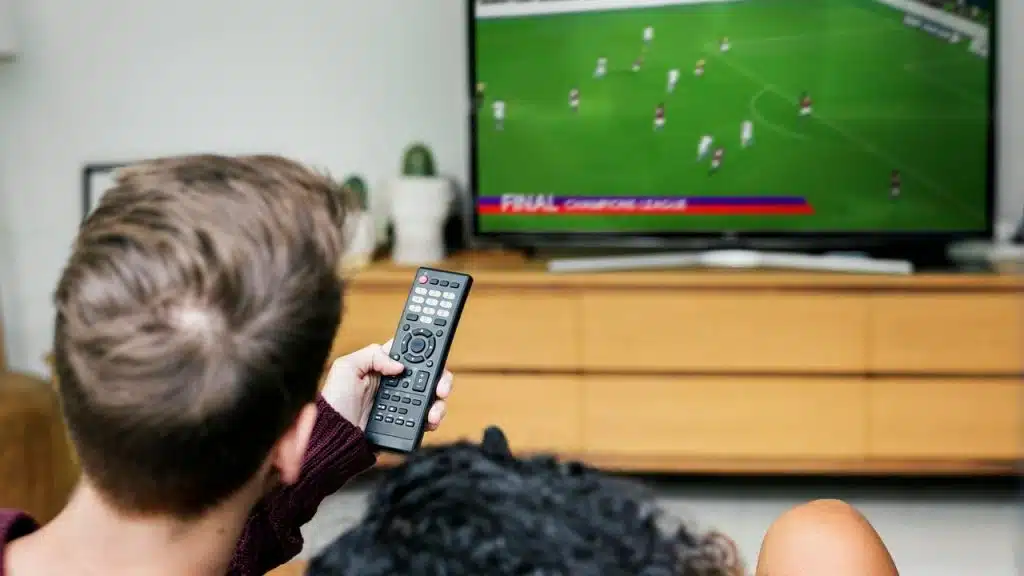 A person holding a TV remote while watching a soccer match on screen.