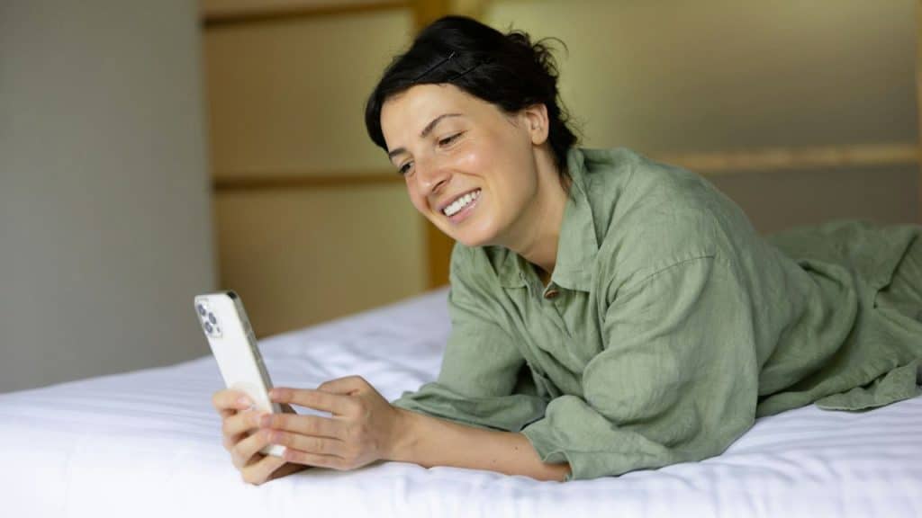 A smiling woman in green pajamas lies on a bed while looking at her phone.