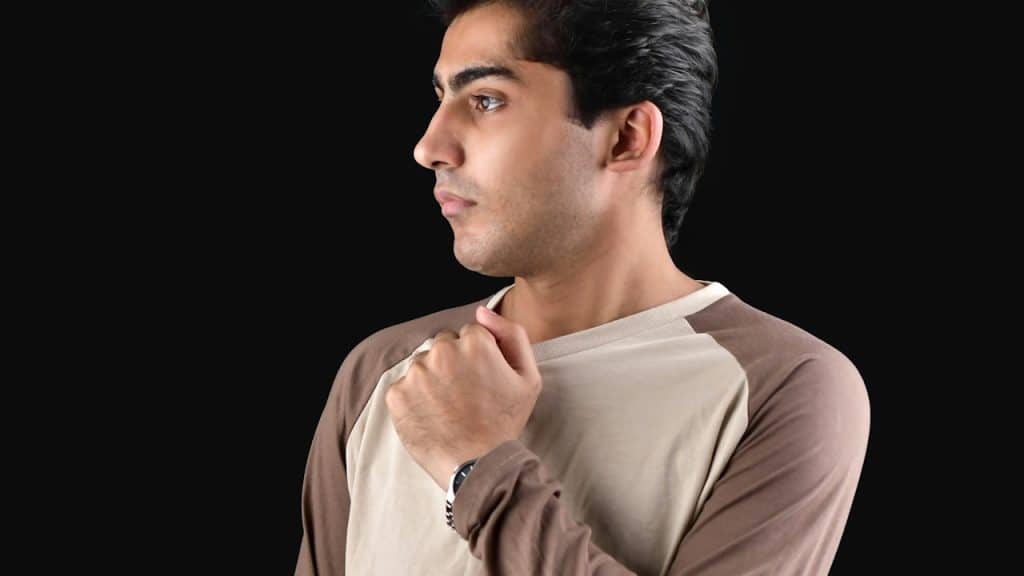 A man wearing a raglan sleeve shirt and holding it by the front. 