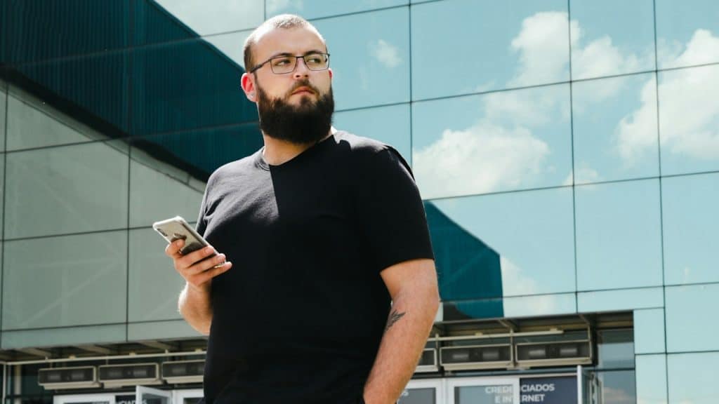 A bearded man wearing glasses and holding his phone stands outdoors.