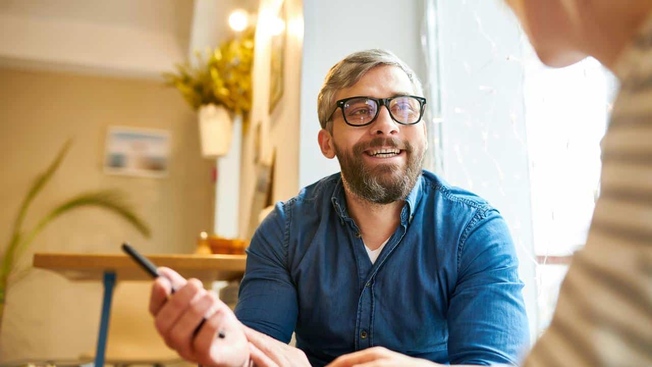A bearded man in glasses holding a pen and smiling indoors.
