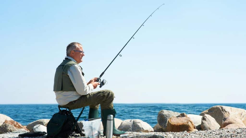 A man fishes while sitting on a rocky shore.