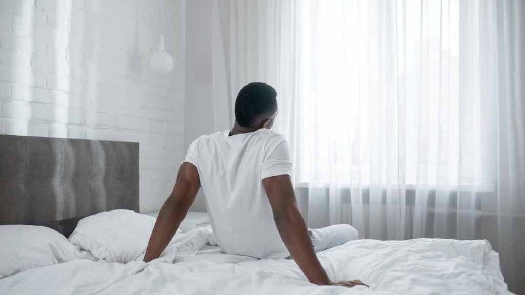 A man sitting on a bed looking out a bright window.