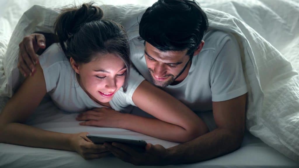 A couple lying in bed under a blanket, looking at a tablet together.