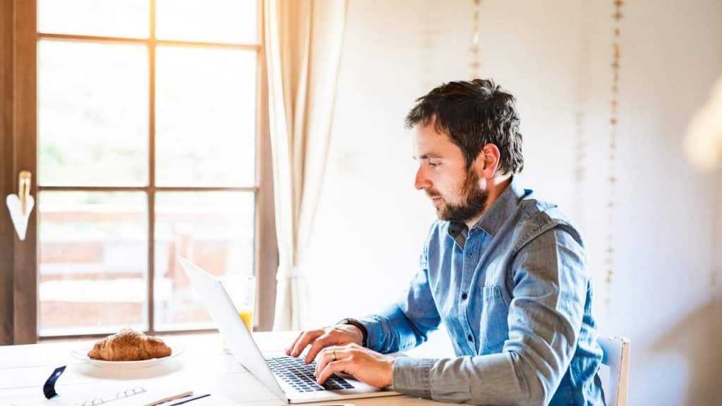 A man working on a laptop at a sunlit table with breakfast nearby.