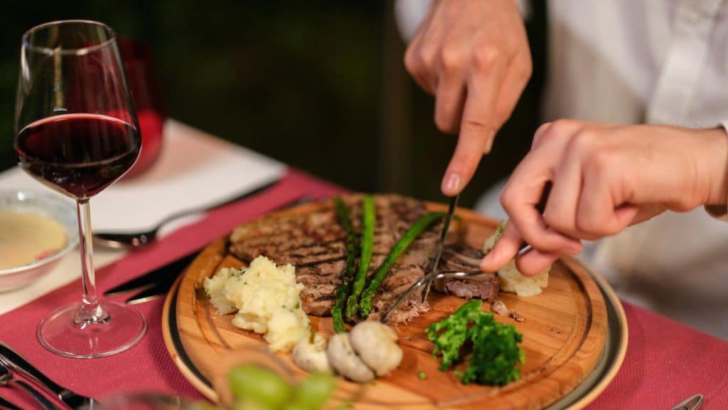 A person cutting steak with wine and sides.