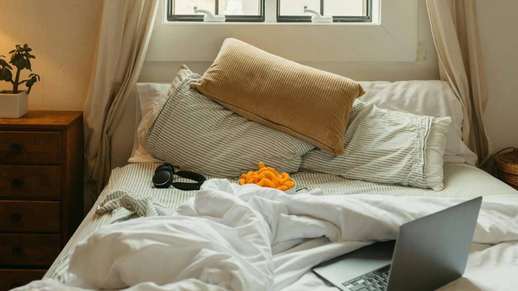 A messy bed with pillows, snacks, headphones, and a laptop.