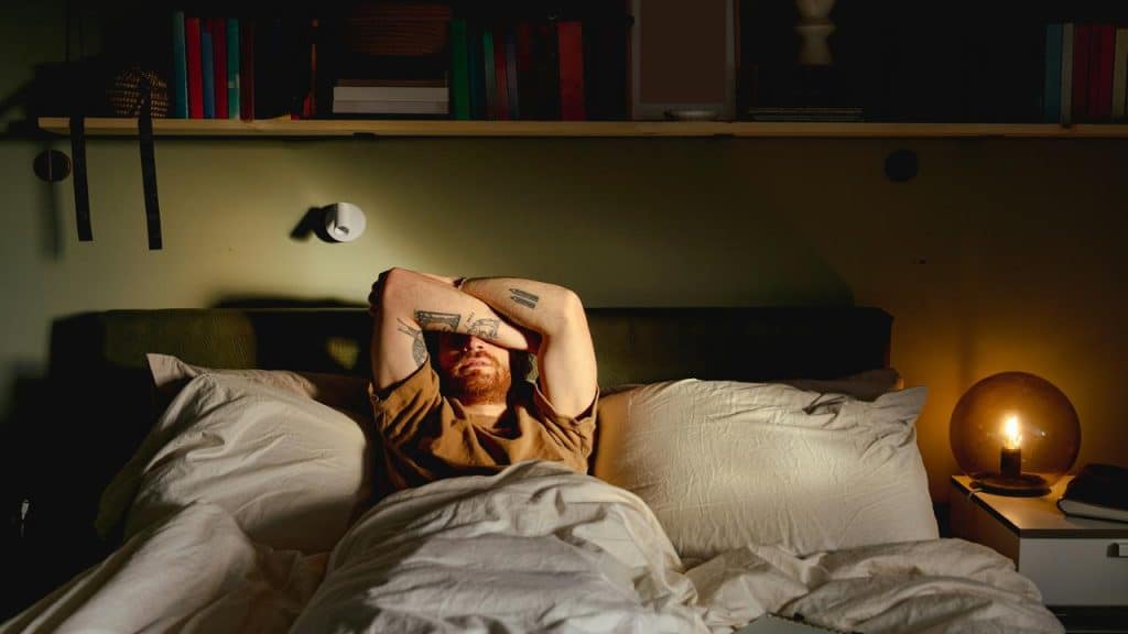 A man lying in bed with his arms over his face in dim light.