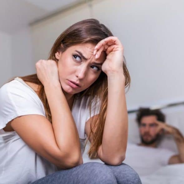 Woman sitting on the edge of the bed, deep in thought, while partner looks unaware.