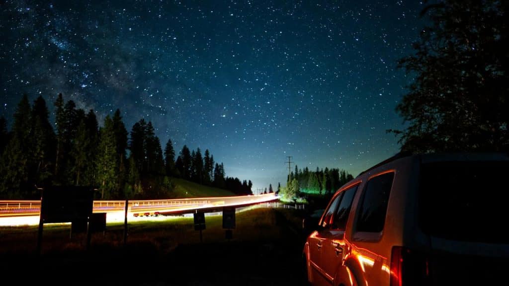 A car parked under a starry night sky by a forest road.