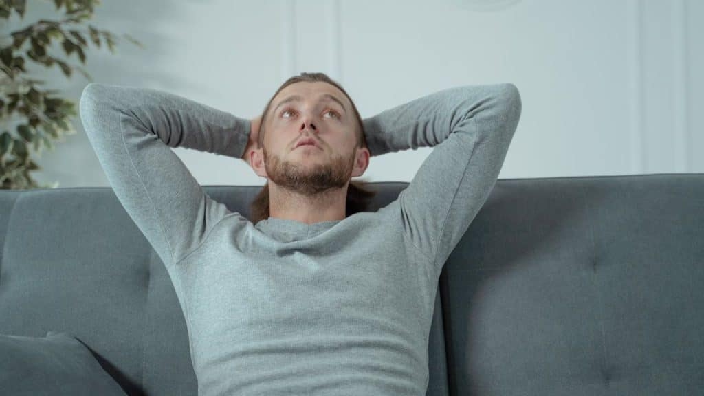 A man with a beard and long hair leans back on a gray couch, hands behind his head.