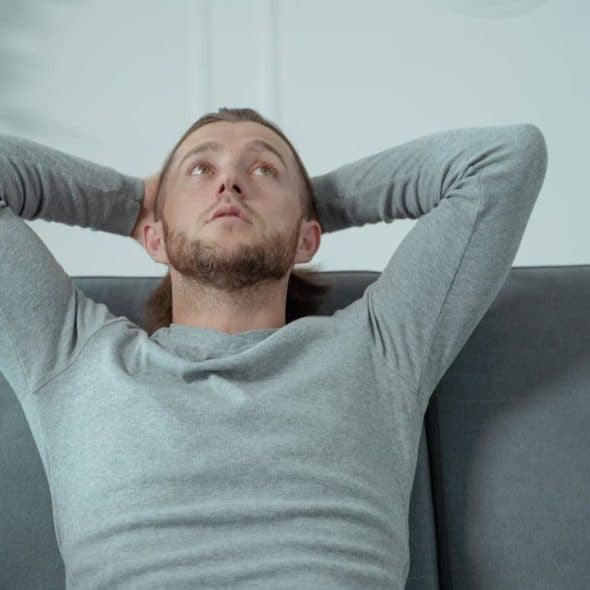 A man with a beard and long hair leans back on a gray couch, hands behind his head.
