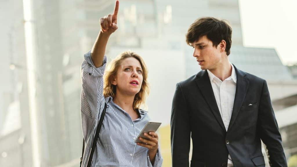 A woman points upward while holding a phone, as a man in a suit looks at her.