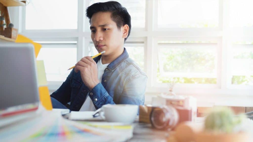 A man in a denim jacket sits at a desk with a laptop, holding a pencil to his chin.