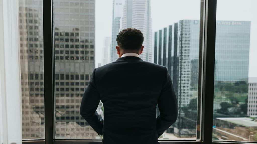 A man in a dark suit stands with his back to the camera, looking out a window at city buildings.
