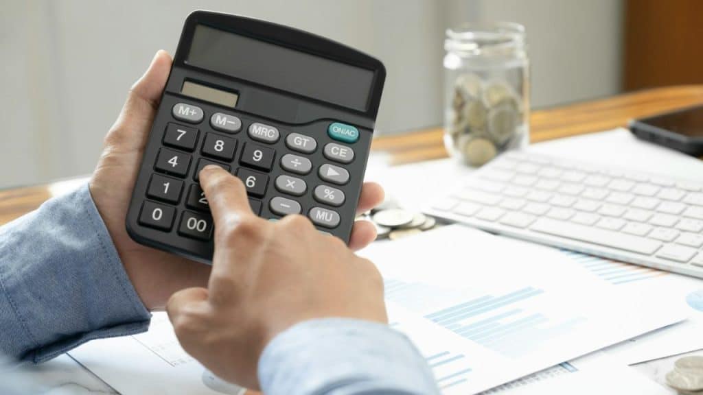 A person's hands are shown using a calculator, with papers, coins, and a keyboard on a desk.