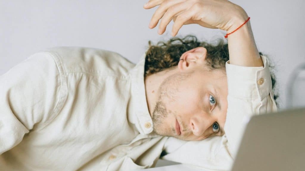 A man with curly brown hair and a beard rests his head on a white desk.