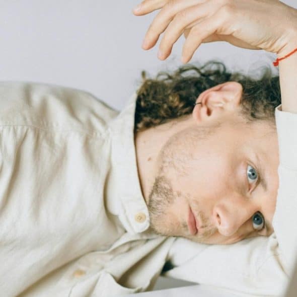 A man with curly brown hair and a beard rests his head on a white desk.
