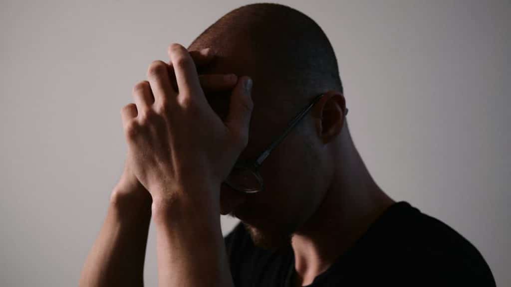 A man with short hair and glasses has his hands covering his face in a dimly lit setting.