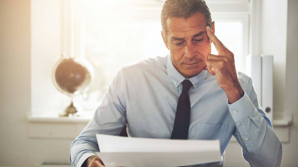 A man in a light blue shirt and tie looks intently at papers, with his hand on his temple.