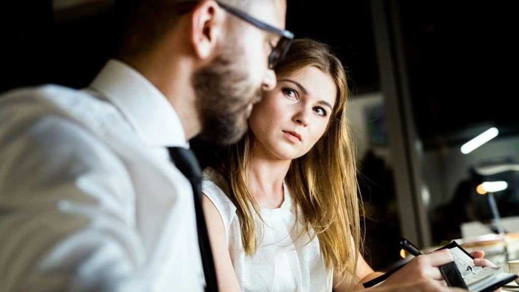 A woman with long hair looks intently at a man wearing glasses and a tie, holding a notebook.