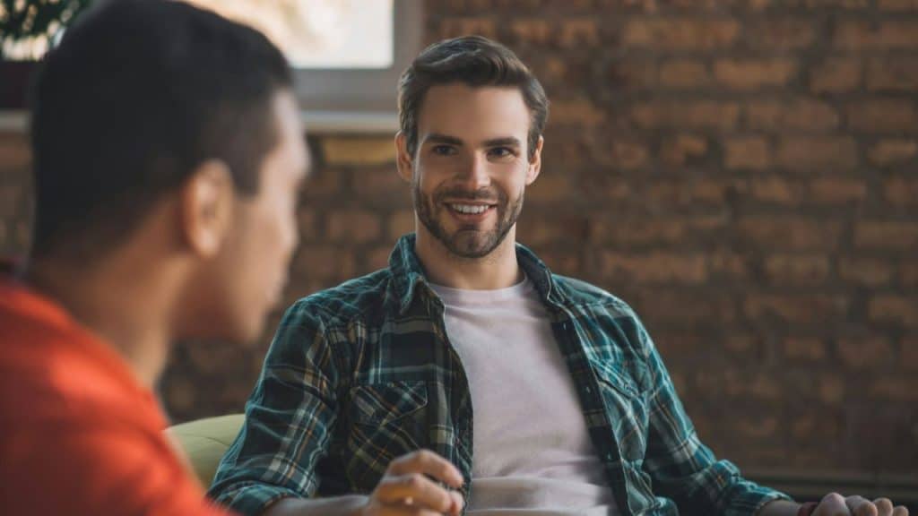 A smiling man in a plaid shirt looks towards another person with a brick wall in the background.