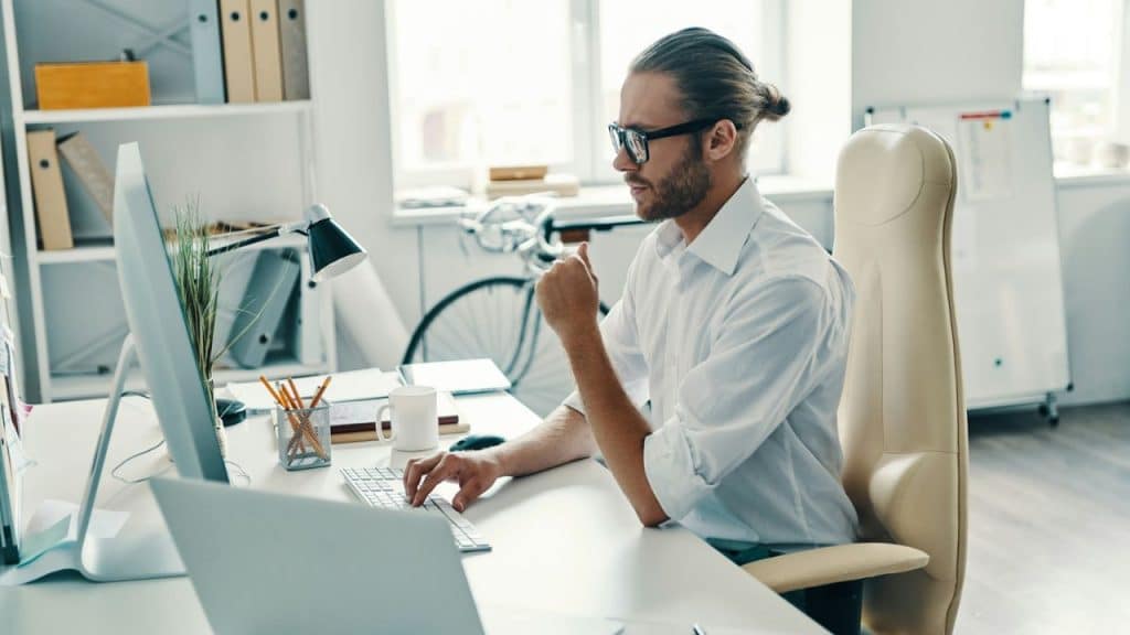 A man with a bun and glasses sits at a desk, typing on a keyboard with two monitors.