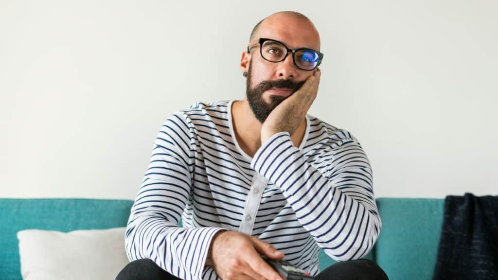 A man with a beard and glasses sits on a couch, holding a remote and resting his head on his hand.