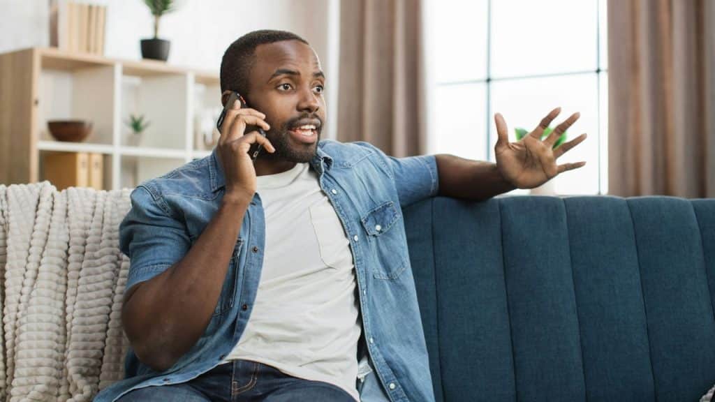 A man on a couch talks on the phone, gesturing with his hand.