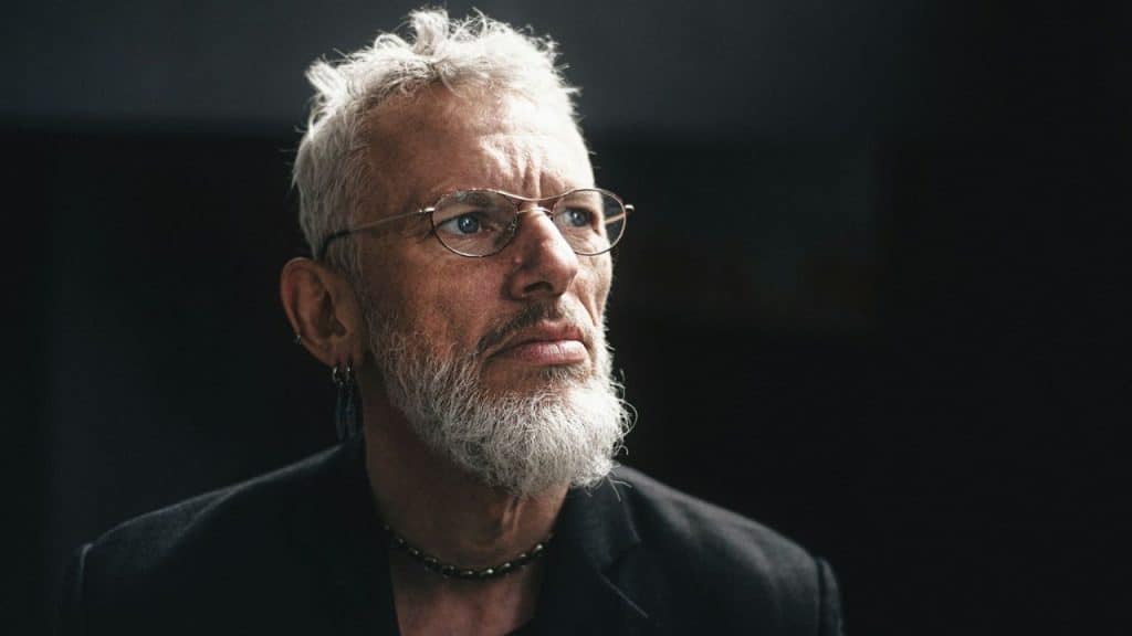 A middle-aged man with gray hair and a beard, wearing glasses, looks up thoughtfully.