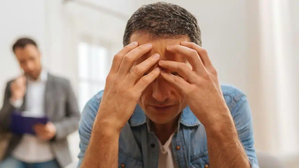 A man in a denim shirt sits with his head in his hands, with another man visible in the background.
