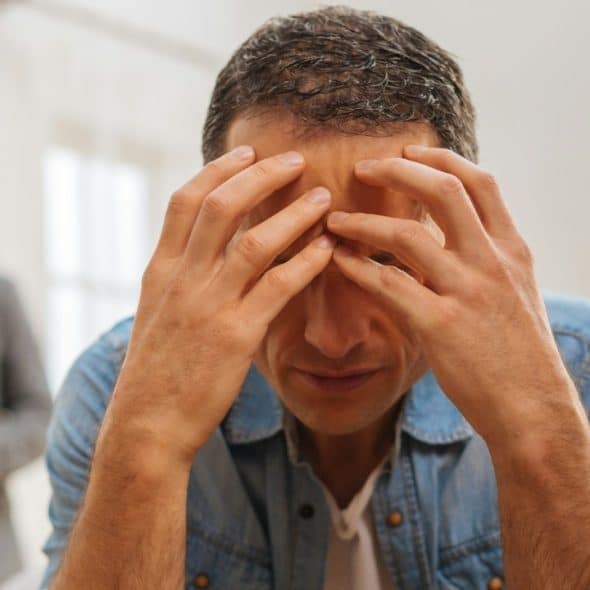 A man in a denim shirt sits with his head in his hands, with another man visible in the background.