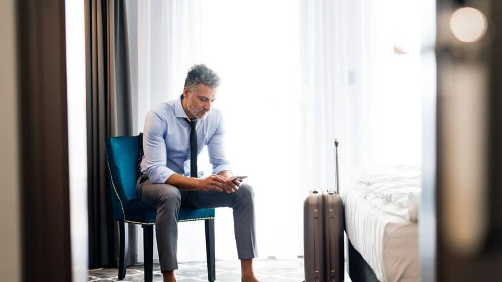 A man in a dress shirt and tie sits barefoot on a blue chair, looking at his phone beside a suitcase.