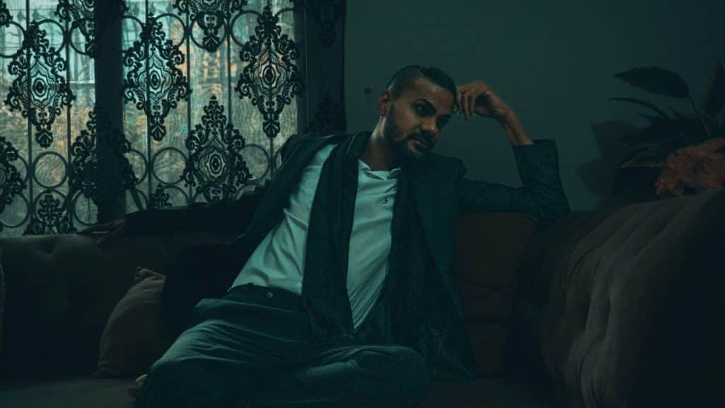 A man in a suit jacket and white shirt reclines on a couch, looking pensive.
