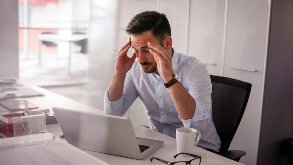 A man in a blue shirt clutches his head, looking stressed at a laptop in an office.