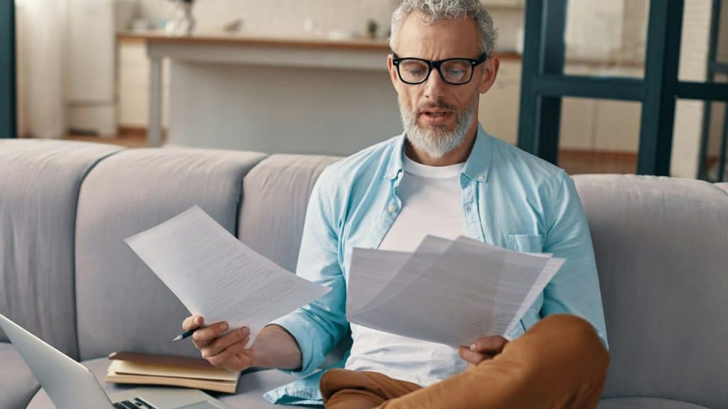 A man with glasses and a beard sits on a couch, intently reading papers.