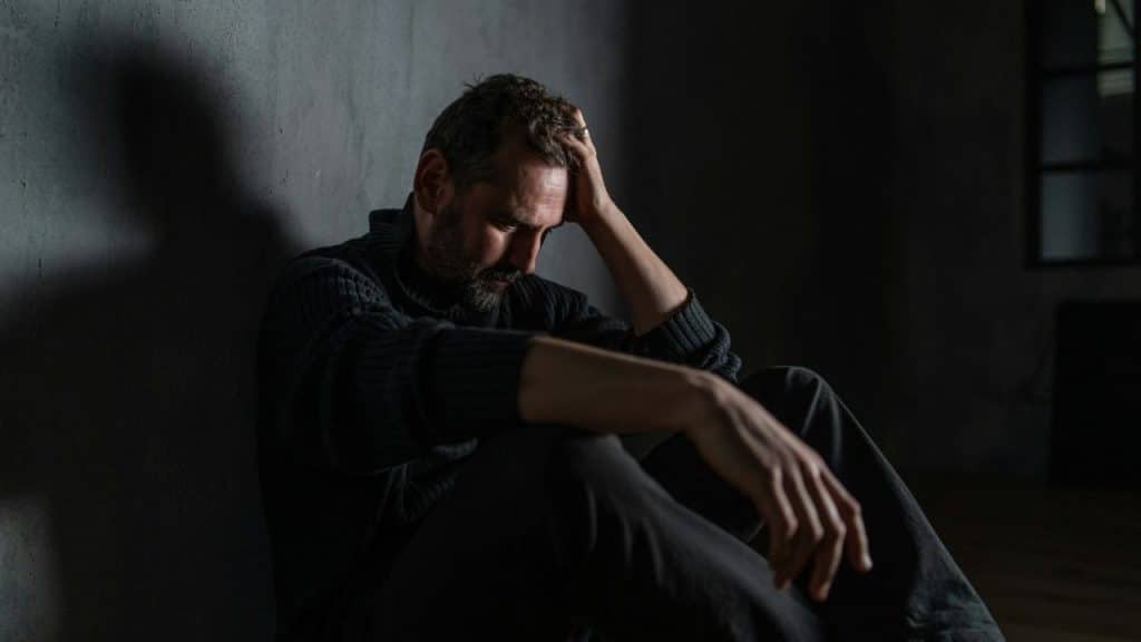 A depressed man with a beard sits on the floor with his hand on his head.