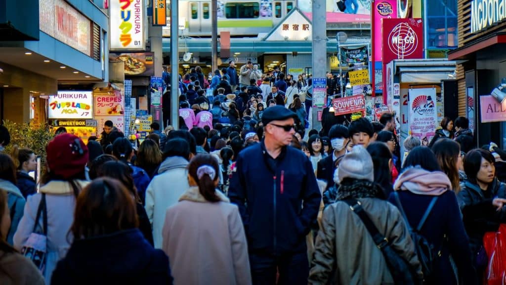 A man with sunglasses and a black cap stares forward amidst a bustling street crowd in Japan.