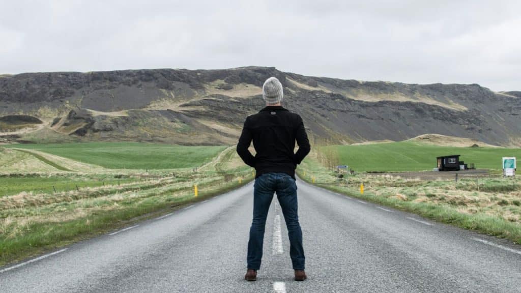A man in a black jacket and jeans stands in the middle of a road, looking at hills.