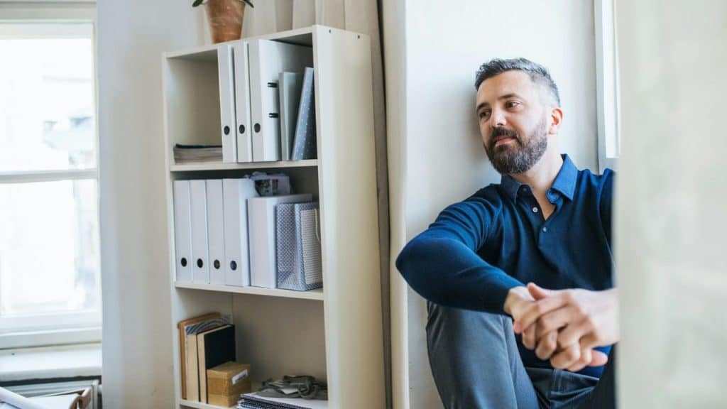 A man with a beard sits on the floor leaning against a wall, looking thoughtfully into the distance.
