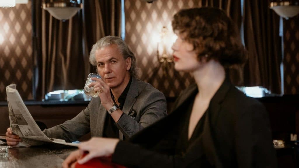 A man with long gray hair and a newspaper sips a drink at a bar.