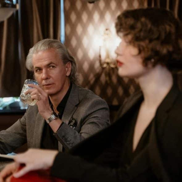 A man with long gray hair and a newspaper sips a drink at a bar.