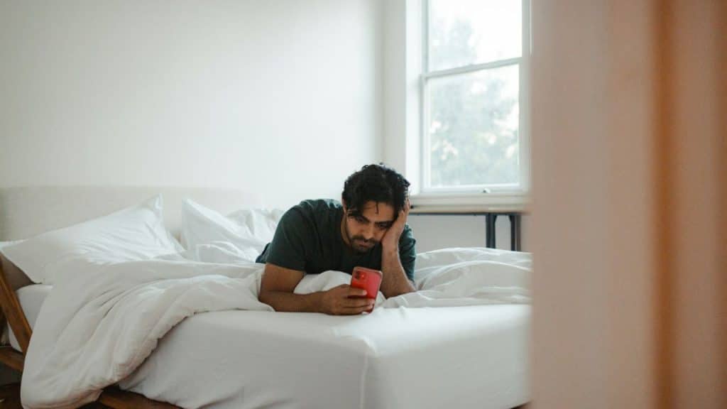A man with dark hair and a beard lies in bed, looking at a red smartphone.
