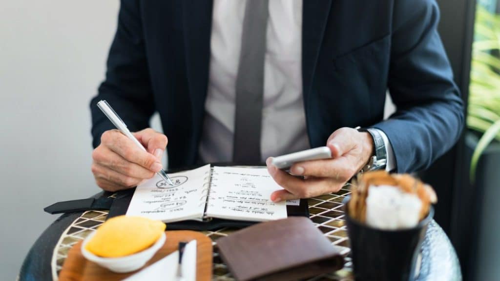 A man in a suit holds a pen and phone, writing in a notebook on a table.