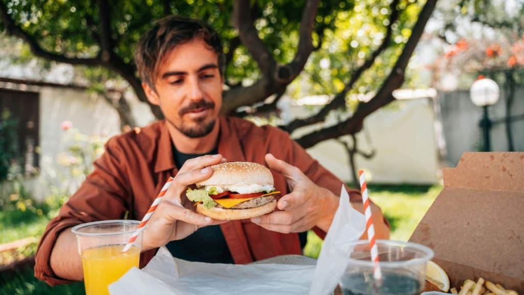 A man outdoors holds a hamburger, with drinks and food on the table.