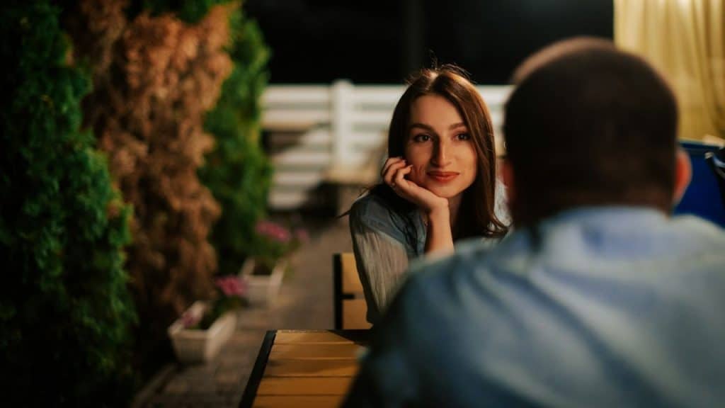 A woman with long dark hair smiles at a man across a table, resting her chin on her hand.