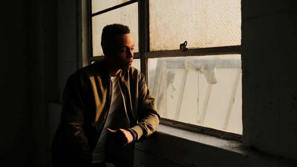 A person in a bomber jacket looks out a window, illuminated by sunlight.